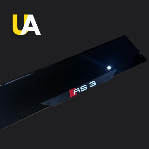 Close-up of a glossy, reflective black trim or panel with a badge for 'RS 3' in white and red, likely a custom door sill for an Audi car. The 'UA' logo in yellow and white is visible in the top left corner.
