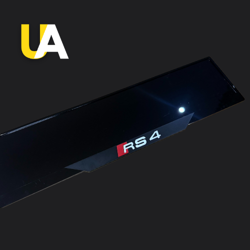 Close-up of a glossy, reflective black trim or panel with a badge for 'RS 4' in white and red, likely a custom door sill for an Audi car. The 'UA' logo in yellow and white is visible in the top left corner.