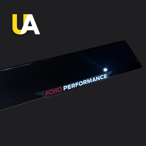 Close-up of a glossy, reflective black trim or panel with the text 'FORD' in red and 'PERFORMANCE' in white, likely a custom door sill or trim piece. The 'UA' logo in yellow and white is visible in the top left corner.
