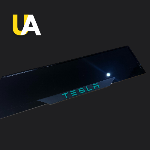 Close-up of a glossy, reflective black trim or panel with the word 'TESLA' illuminated in blue-green light, likely a custom door sill or trim piece. The 'UA' logo in yellow and white is in the top left corner.