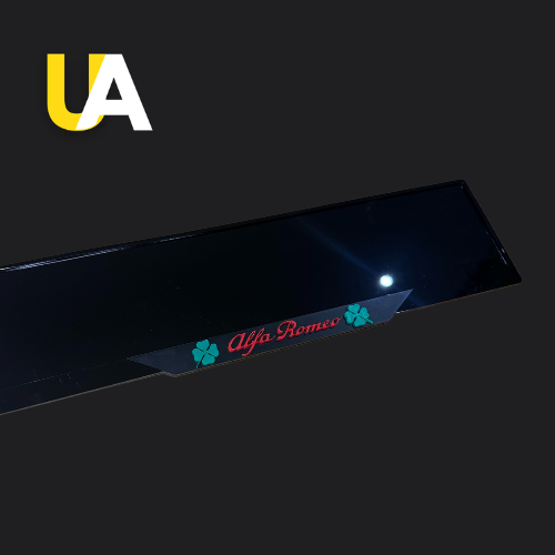 Close-up of a glossy, reflective black trim or panel featuring a small decal or badge with the text 'Alfa Romeo' in red and two green four-leaf clover symbols (Quadrifoglio). The 'UA' logo in yellow and white is in the top left corner.
