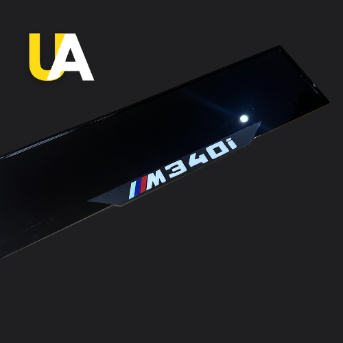 Close-up of a glossy, reflective black trim or panel with the text 'M340i' in white, preceded by the BMW M stripes (blue, dark blue, and red), likely a custom door sill. The 'UA' logo in yellow and white is visible in the top left corner.