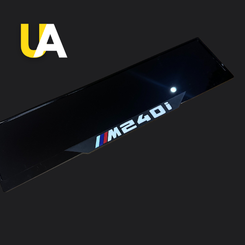 Close-up of a glossy, reflective black trim or panel with the text 'M340i' in white, preceded by the BMW M stripes (blue, dark blue, and red), likely a custom door sill. The 'UA' logo in yellow and white is visible in the top left corner.