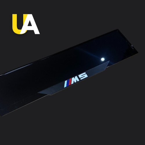 Close-up of a glossy, reflective black trim or panel with the text 'M5' in white, preceded by the BMW M stripes (blue, dark blue, and red), likely a custom door sill. The 'UA' logo in yellow and white is visible in the top left corner.