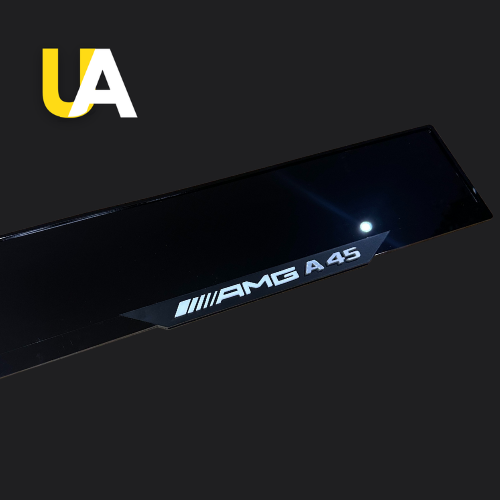 Close-up of a glossy, reflective black trim or panel with the white text 'AMG A 45,' preceded by the four white parallel lines logo, likely a custom door sill for a Mercedes-Benz car. The 'UA' logo in yellow and white is visible in the top left corner.