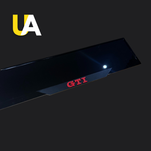 Close-up of a glossy, reflective black trim or panel with the red text 'GTI,' likely a custom door sill or trim piece for a Volkswagen car. The 'UA' logo in yellow and white is visible in the top left corner.